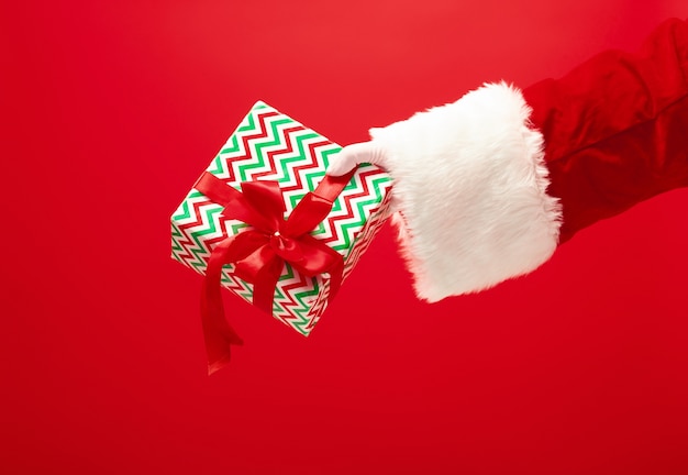 Free Photo the hand of santa claus holding a gift on red