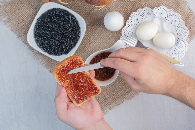 Hand slathers jam on bread with knife.