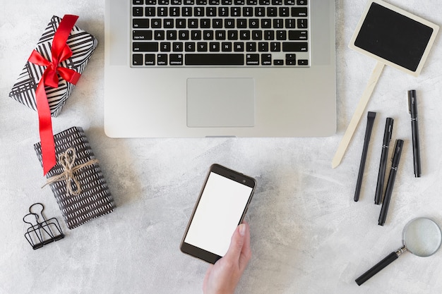 Hand with smartphone near laptop, present boxes and stationery