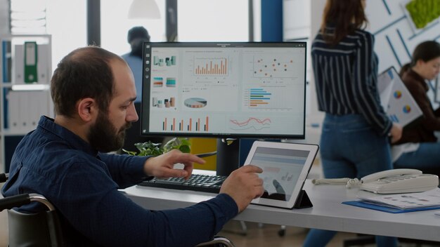 Handicapped invalid man employee sitting immobilized in wheelchair working at notepad and computer in same time analysing financial graphs, pc showing data prosessing for project regarding economy