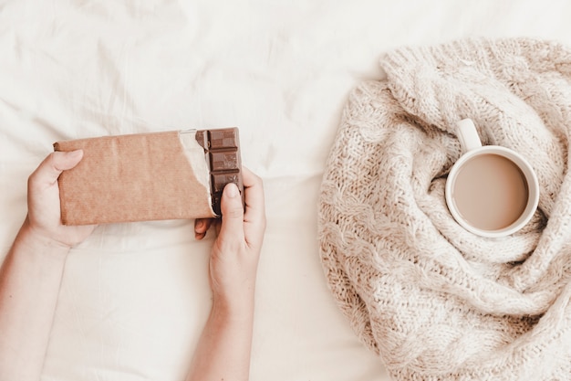 Hands holding chocolate near cup of hot drink in plaid on bedsheet