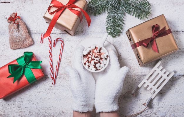 Hands holding a cup with marshmallows and christmas gifts