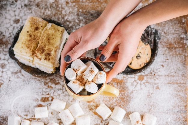 Hands holding cup with marshmallows near sweets plate