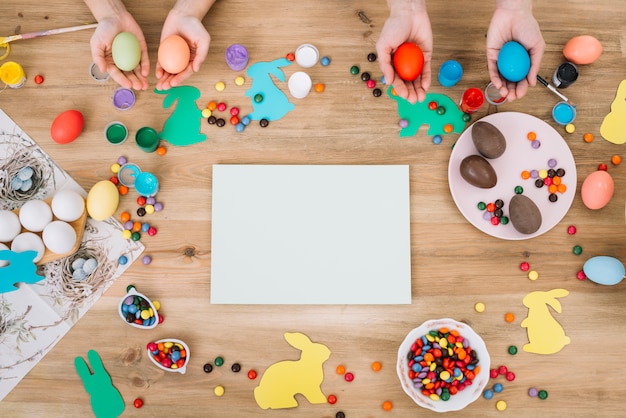 Hands holding easter eggs with colorful candies and white paper over the wooden table