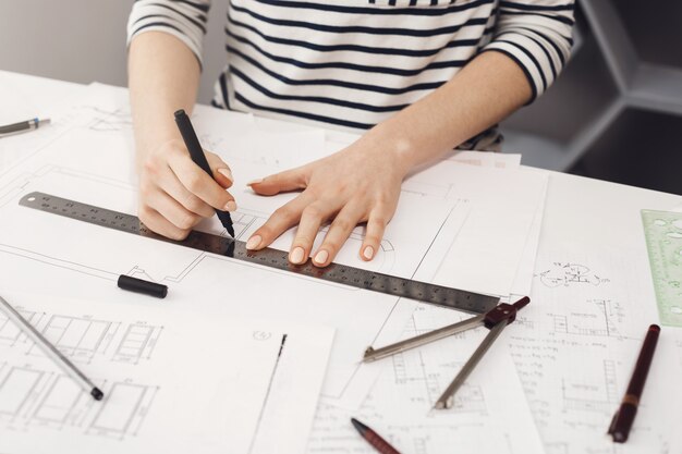 Hands of successful young female architect in striped shirt sitting at white table in home, making drawings with pen and ruler, doing project of her future room.