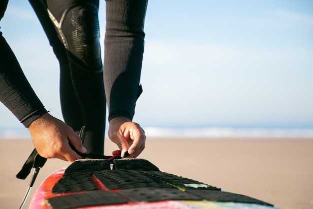 Free photo hands of surfer in wetsuit tying surfboard to his ankle on ocean beach