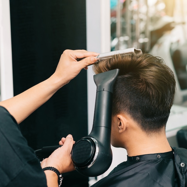 Hands of unrecognizable hairdresser giving male customer blowdry with hairdryer and comb