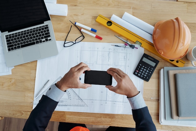 Hands of unrecognizable man holding smartphone over technical drawing