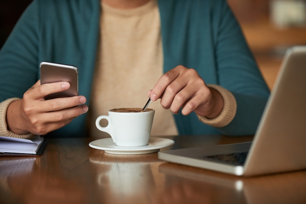 Hands of unrecognizable man sitting in cafe, holding smartphone and stirring coffee