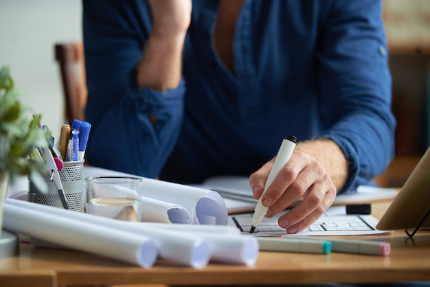 Hands of unrecognizable man sitting at desk in office and drawing on floor plan with marker