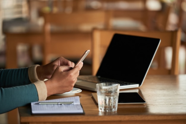 Hands of unrecognizable man using smartphone in cafe, and laptop on table