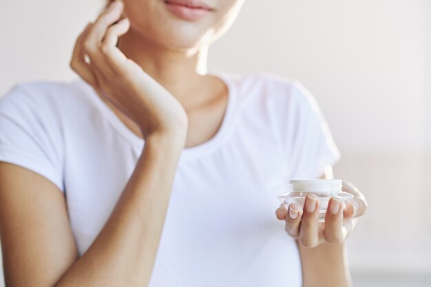 Hands of unrecognizable woman holding pot and spreading face cream on skin 