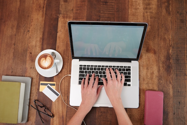 Free Photo hands of unrecognizable woman working on laptop, with cappuccino and credit cards on table