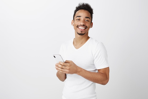 Handsome african american guy listening music in wireless earphones and holding mobile phone, smiling pleased