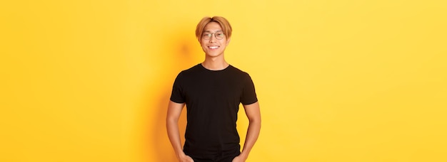 Free photo handsome asian blond guy in glasses standing in black clothes and smiling pleased yellow background