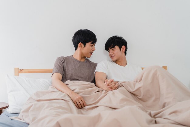 Handsome Asian gay couple talking on bed at home. Young Asian LGBTQ+ guy happy relax rest together spend romantic time after wake up in bedroom at modern house in the morning .