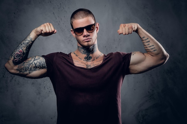 Free photo handsome, athletic male in sunglasses with tattoos on his arms and neck posing over grey artistic background in a studio.