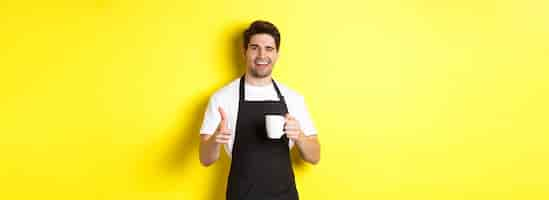 Free photo handsome barista in black apron holding coffee cup pointing finger at you inviting visit cafe standi