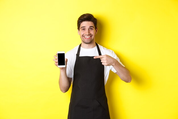 Handsome barista in black apron pointing finger at mobile screen, showing app and smiling, standing over yellow background.