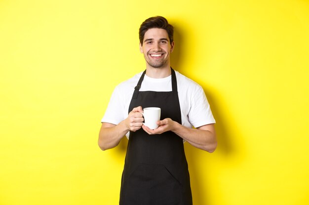 Handsome barista serving coffee, bring cup, standing in black apron with friendly smile.