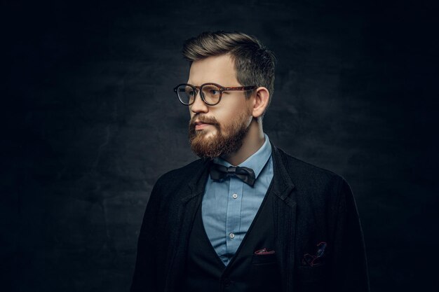 Handsome bearded elegant male in eyeglasses dressed in a dark blue suit with bow tie over grey vignette background.