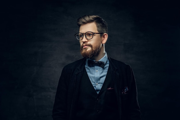 Handsome bearded elegant male in eyeglasses dressed in a dark blue suit with bow tie over grey vignette background.