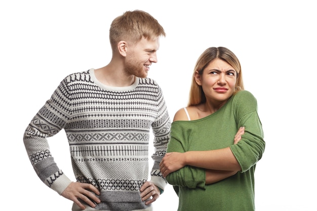 Handsome bearded guy dressed in sweater smiling and looking at attractive woman who standing in closed posture with arms crossed, feeling confused as she doesn't like or understand his silly joke