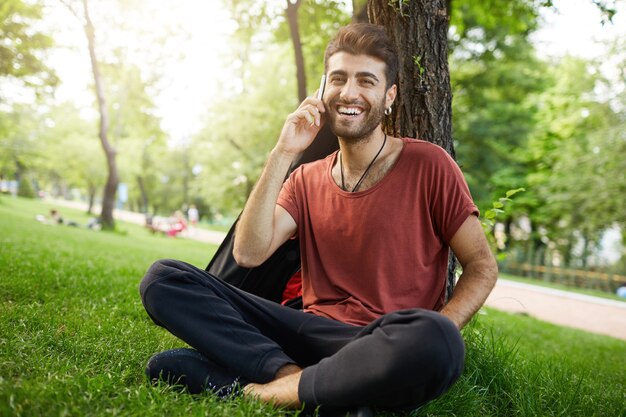 Handsome bearded guy resting in park on grass, talking on mobile phone and smiling happy