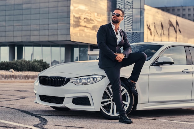 Handsome bearded male in sunglasses dressed in a black suit sitting on a luxury car against a skyscraper.