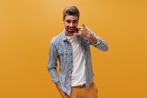 Handsome bearded man in bright pants white tshirt and checkered shirt smiles looks into camera and shows call sign on orange background