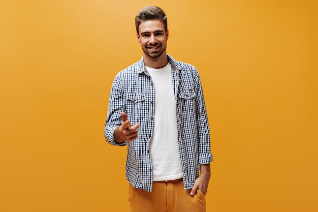 Free Photo handsome bearded man in orange pants white tshirt and checkered shirt smiles sincerely and points at camera on orange background