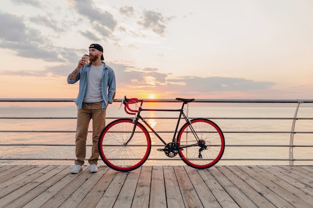 Handsome bearded man traveling with bicycle in morning sunrise by the sea drinking coffee, healthy active lifestyle traveler