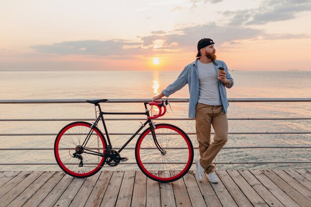 Handsome bearded man traveling with bicycle in morning sunrise by the sea drinking coffee, healthy active lifestyle traveler