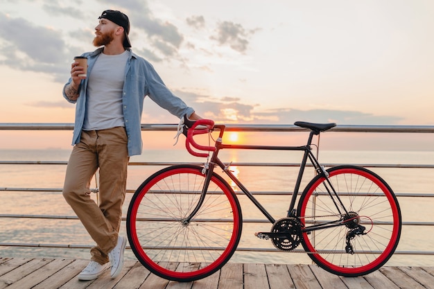 Free photo handsome bearded man traveling with bicycle in morning sunrise by the sea drinking coffee, healthy active lifestyle traveler