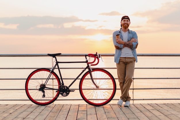 Handsome bearded man traveling with bicycle in morning sunrise by the sea drinking coffee, healthy active lifestyle traveler