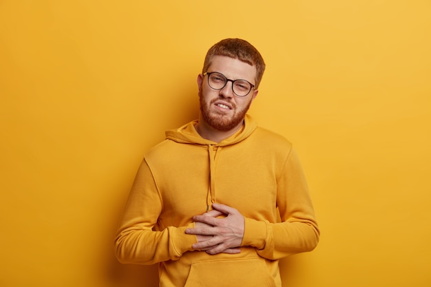 Free Photo handsome bearded man with ginger hair feels stomachache, suffers from indigestion and abdomainal cramps after eating spoiled product, wears glasses and sweatshirt, isolated on yellow wall