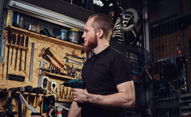 A handsome bearded redhead worker with haircut,  holding an electric screwdriver, working in a repair shop.