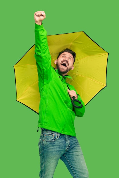 Handsome bearded smiling happy young man holding umbrella and looking at camera isolated on vivid trendy green studio.