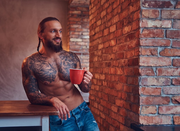 Free Photo handsome bearded tattoed male with a stylish haircut and muscular body, drinks coffee, leaning on a table.