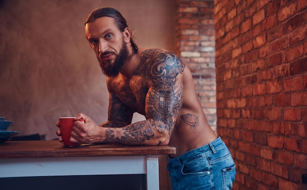 Handsome bearded tattoed male with a stylish haircut and muscular body, drinks coffee, leaning on a table.