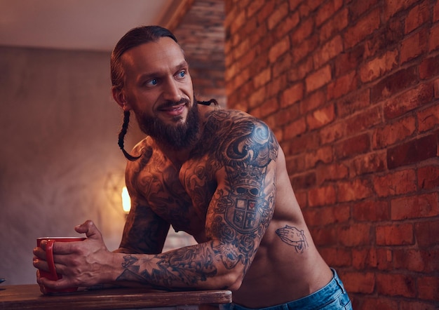 Free photo handsome bearded tattoed male with a stylish haircut and muscular body, drinks coffee, leaning on a table.