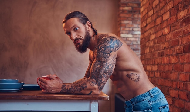 Free Photo handsome bearded tattoed male with a stylish haircut and muscular body, drinks coffee, leaning on a table.