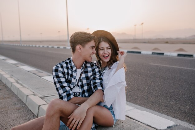 Handsome boy in checkered shirt touching his girlfriend's knee and laughing, while sitting on the road. Portrait of smiling loving couple spending time together on the highway enjoys amazing sunset