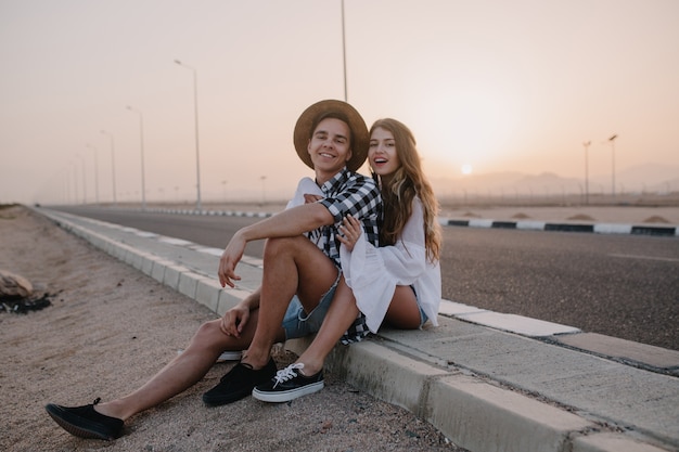 Handsome boy wearing hat and woman in white vintage blouse sitting together on the road and enjoys beauiful sunset. Charming long-haired young woman resting near the highway with her boyfriend