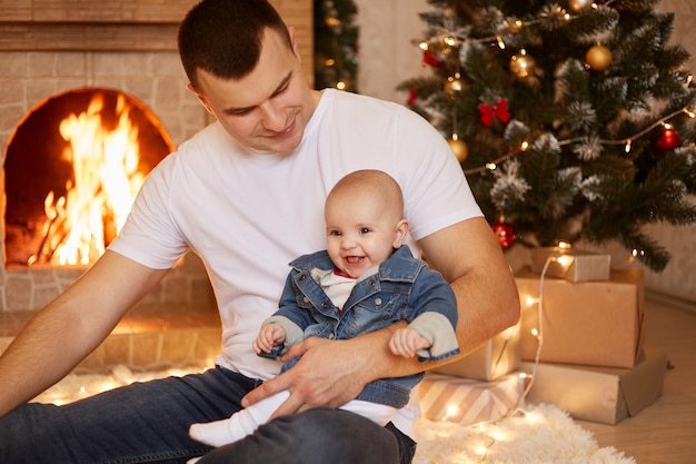 Free photo handsome brunet father holding his small infant daughter in hands, looking at his cute charming kid, sitting near fireplace and xmas tree at home, happy new year.