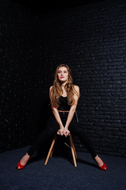 Free photo handsome brunette girl wear on black and red high heels sitting and posing on chair at studio against dark brick wall studio model portrait