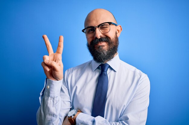 Handsome business bald man with beard wearing elegant tie and glasses over blue background smiling with happy face winking at the camera doing victory sign Number two