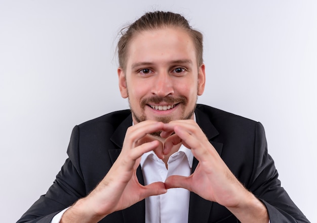 Handsome business man wearing suit aking heart gesture with fingers looking at cmera with smile on face standing over white background