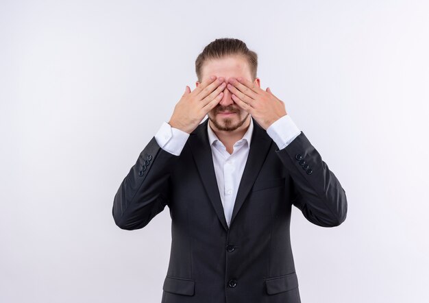 Handsome business man wearing suit covering eyes with hands standing over white background