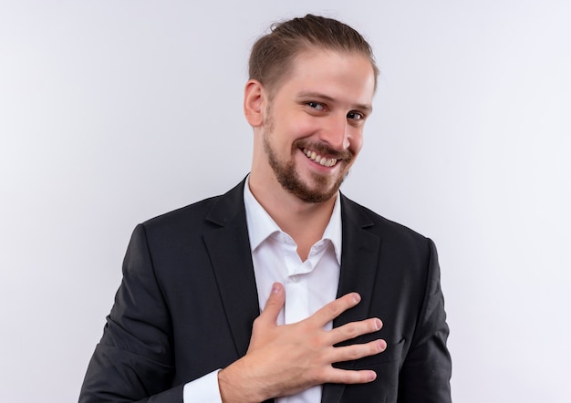 Handsome business man wearing suit holding hand on his chest looking at camera with smile on face standing over white background